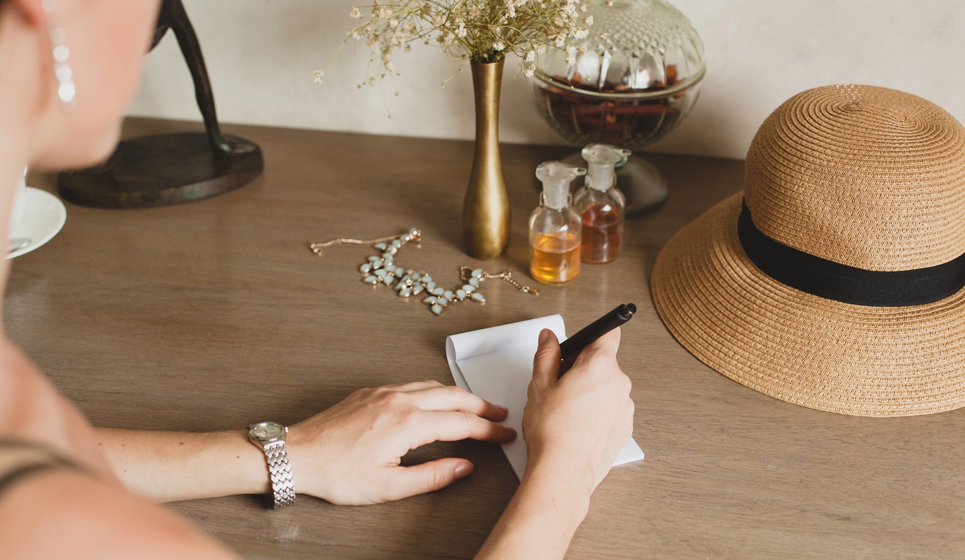 young stylish beautiful woman sitting at table in resort hotel room, writing a letter, holding pen, straw hat, vintage style, hands close-up, details, accessories, travel diary